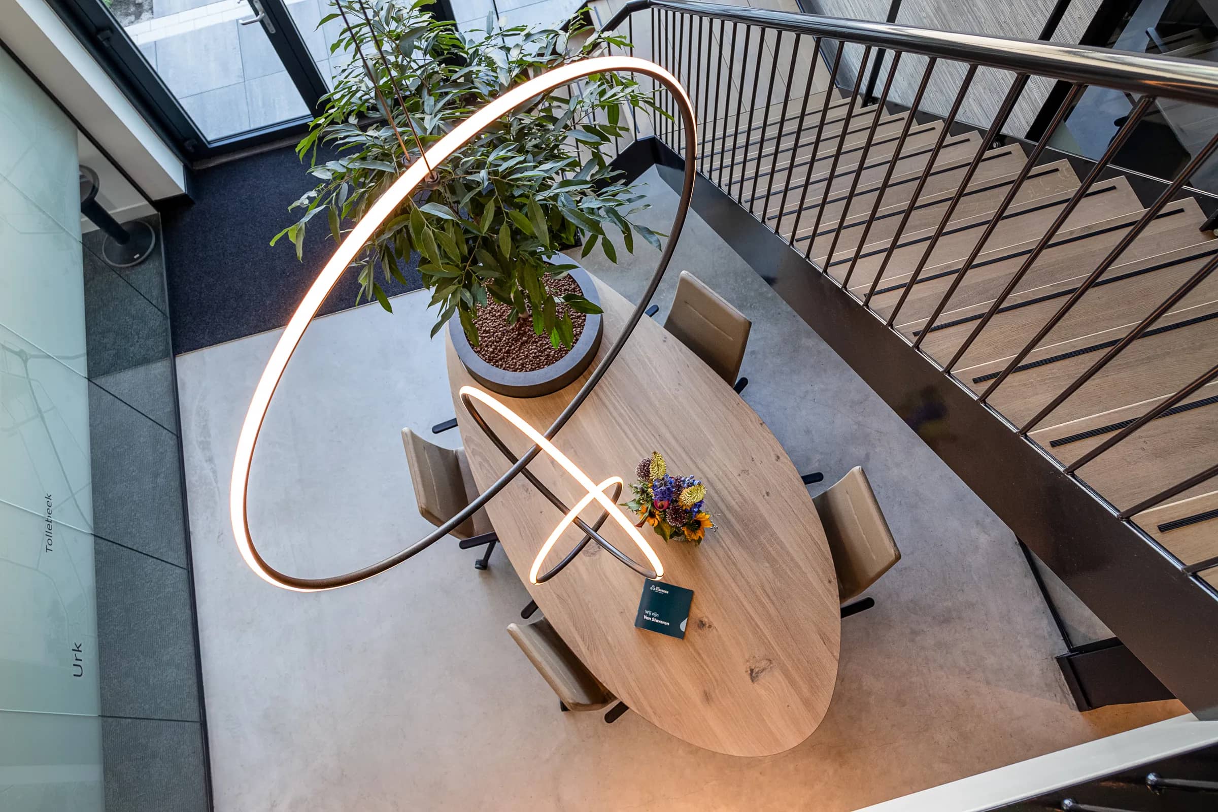Oval oak table seen from above under an LED light ring next to a staircase with a steel railing at Van Staveren